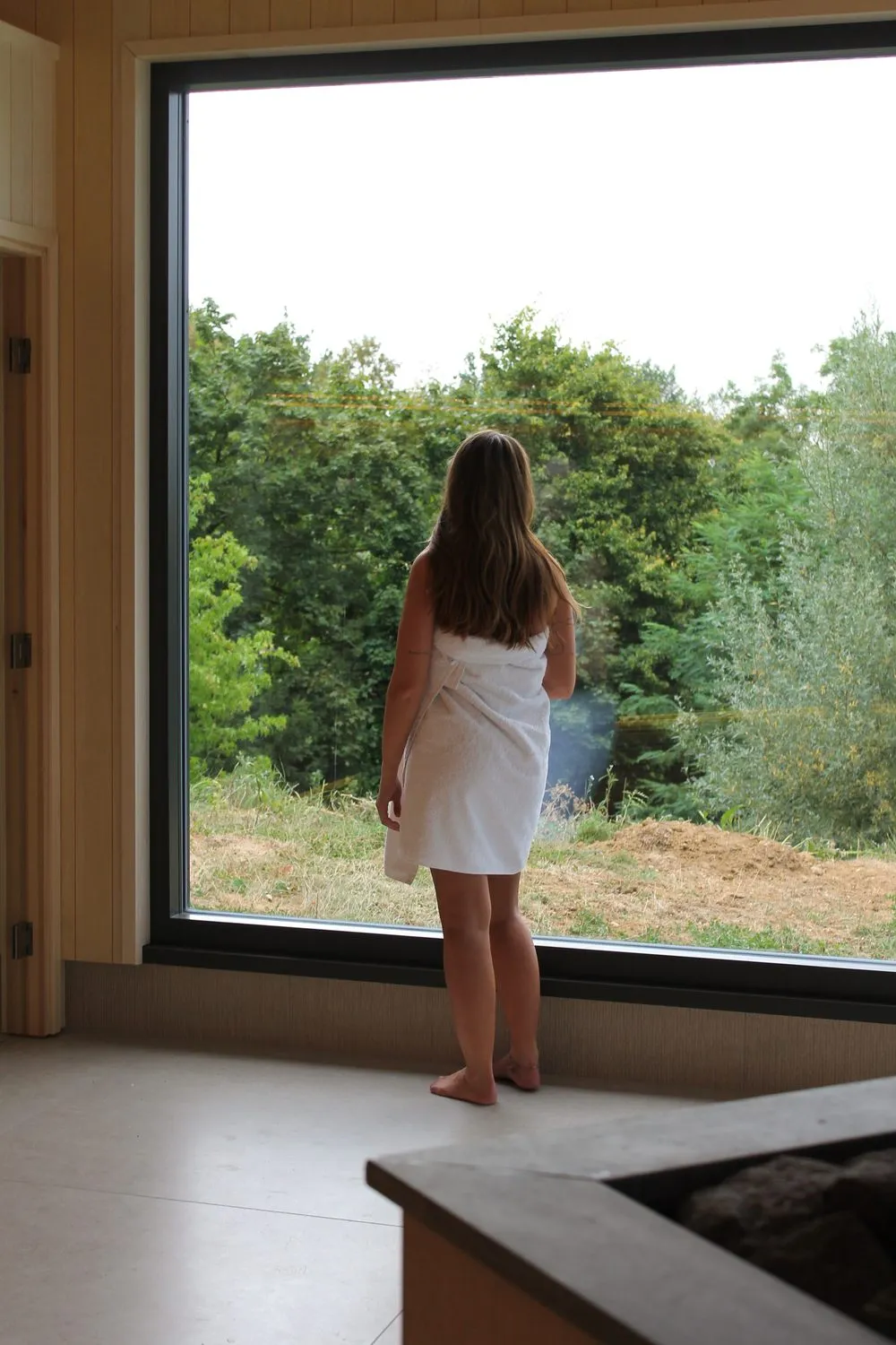 Vrouw in handdoek kijkt door het grote raam van Eir uit over de bomen en het Limburgse heuvelland