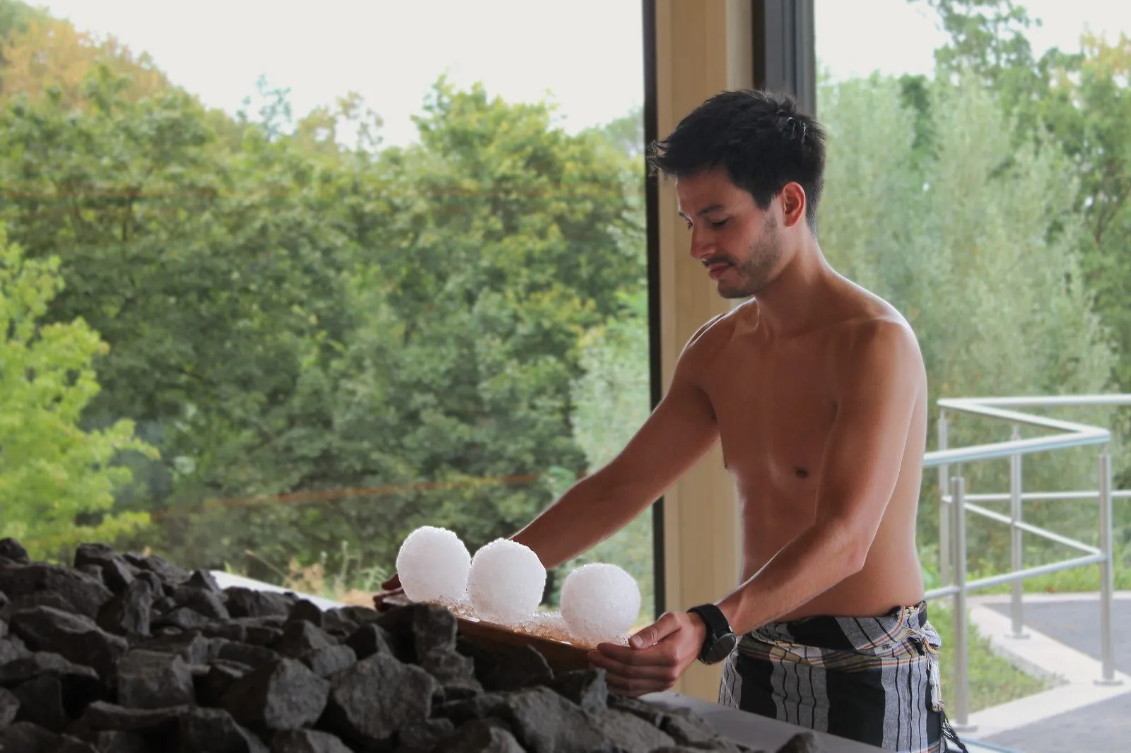 Saunameester legt ijsballen op de kachel van opgietsauna Eir bij Thermae 2000, met door het grote raam uitzicht op de bomen van de Cauberg